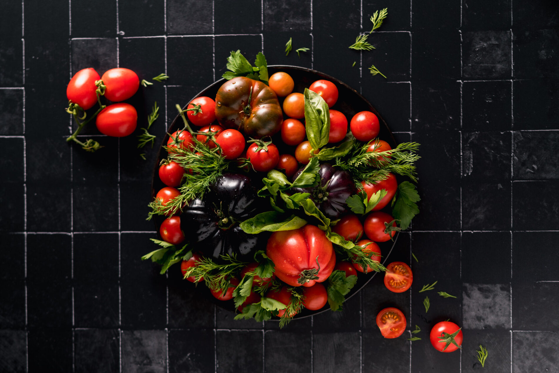 Different red tomatoes arranged on a black tiled floor
